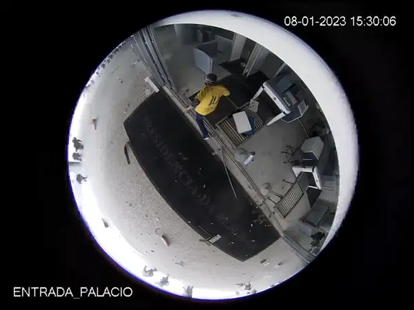 A film still: A fischeye view (a rounded image over a black background), probably from a security camera, with a person with a yellow jacked walking in a vandalized hall. On the bottom, the text Entrada Palacio